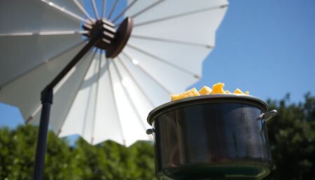 A close-up of a homemade parabolic solar cooker, with a pot of food simmering at its focus, set against a clear blue sky and a backdrop of lush greenery.