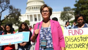 A determined San Diego advocate, JoAnn Fields, stands outside the U.S. Capitol, surrounded by fellow advocates, holding signs that read 'Remember Us' and 'Fund Disaster Recovery'.