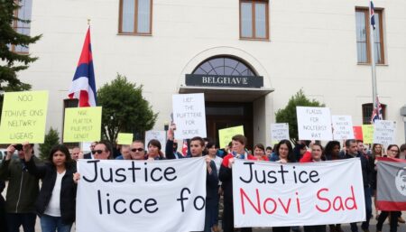 A photo of protesters holding signs and banners in front of the Serbian President's office in Belgrade, with the railway station in Novi Sad in the background, and the words 'Justice for Novi Sad' written on the image.