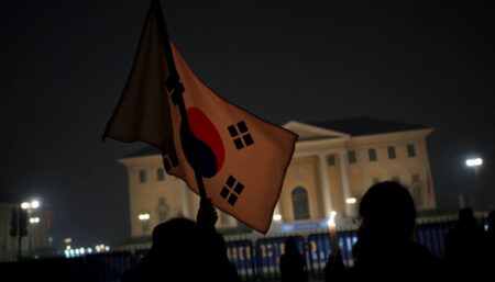 A tense South Korean flag waving in the wind, with a dark silhouette of a protester holding a candle in the foreground, and the presidential office in the background, shrouded in police lights and barricades.