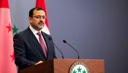 Create an image of Syria's new foreign minister, Asaad Hassan al-Shibani, standing confidently at a podium with the Syrian flag in the background, addressing a press conference. The scene should convey a sense of determination and new beginnings.